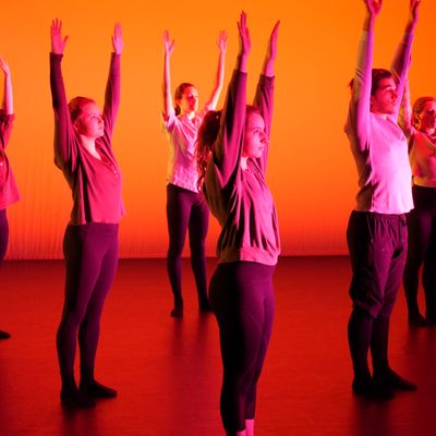 Dancers stand onstage in red-orange light. They uniformly stand still and reach arms to the sky.