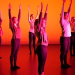 Dancers stand onstage in red-orange light. They uniformly stand still and reach arms to the sky.