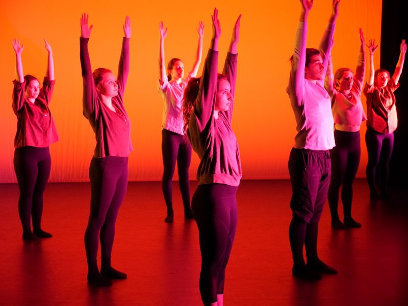 Dancers stand onstage in red-orange light. They uniformly stand still and reach arms to the sky.
