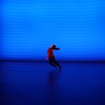 Photo of a dancer captured mid-run. The dancer is alone onstage in blue light.