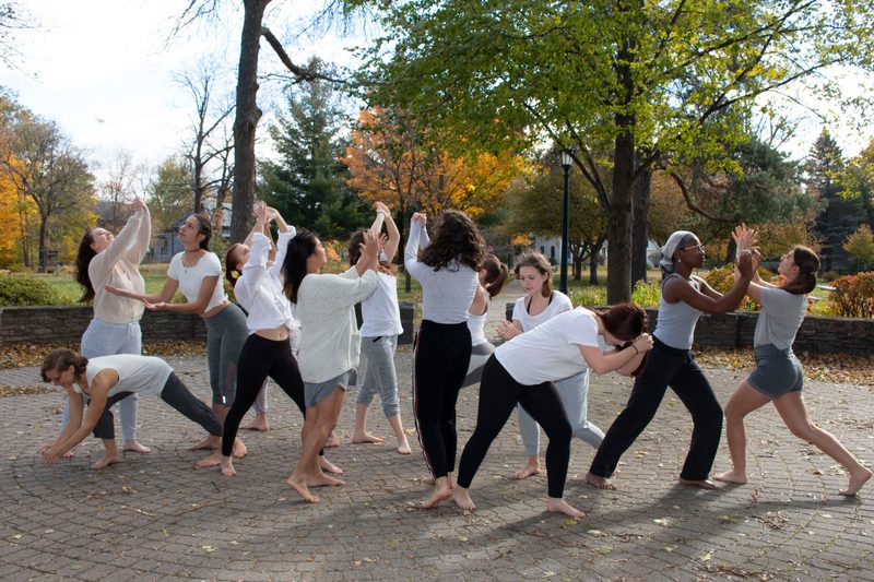 Company members practice a piece outside on a sunny fall day. They are barefoot and striking poses.