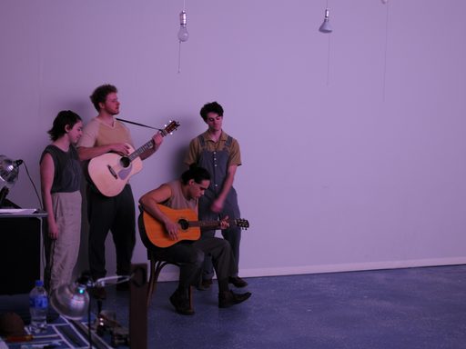 A group of four people in a loose cluster before a purple wall. Two people are playing a guitar.