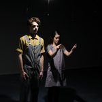 A man and woman stand under a light bulb. The man wears overalls and his hands hang at his sides.