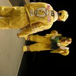 A race car driver in yellow facing a person in a chicken costume