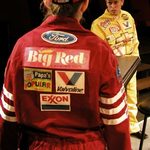 The back of a race car driver in red and another driver in yellow looking at them