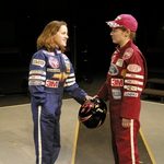 Two women wearing race car driving outfits and holding on to a helmet