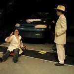 A woman working on a car and a person wearing a white suit standing over her