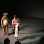 A person in a bear costume walking behind a man without a shirt