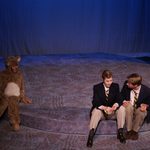 Two men sitting on a step talking, and a person in a bear costume sitting by them