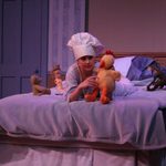 A woman laying in bed wearing a chef hat and playing with a stuffed animal
