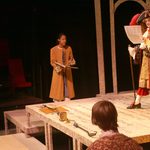 An actor in wealthy red and gold clothing reading from a piece of paper to other actors