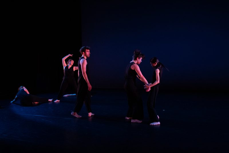 6 dancers perform onstage, in black sleeveless tops and black pants. Four dancers stand, two lay on the floor.