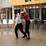 2 dancers perform in Weitz Commons. Both stand. One dancer cradles the head of the second dancer, who is leaning backward.