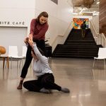 2 dancers perform in Weitz Commons. One dancer sits on the floor with her legs bent at the knees, reaching up and holding hands with the second dancer, who stands.