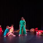 8 dancers in colorful costumes move onstage, some sitting or lying on the floor, two of them standing facing upstage.