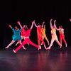 Eight dancers grouped center stage and dressed in bright, colorful costumes jump with arms and legs outstretched.