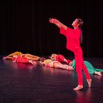 A dancer in red faces stage right, looking to the sky with arms reaching straight upwards. Upstage of her, five dancers in colorful costume roll their full bodies on the floor.
