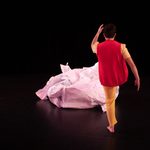 A dancer in yellow pants and a red vest faces upstage, kicking a large, crumpled ball of paper.