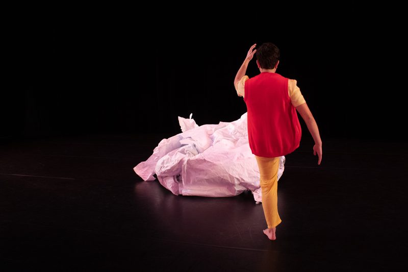 A dancer in yellow pants and a red vest faces upstage, kicking a large, crumpled ball of paper.