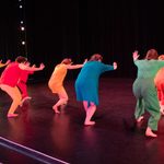 Seven dancers in very bright costumes line up on stage and lunge forward with hands outstreched.