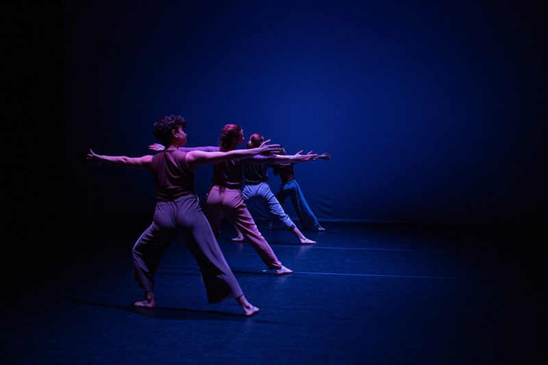 Four dancers are lined up downstage to upstage, with their hands and legs extended, bathed in blue light.