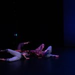 Four dancers form a circle onstage, with their heads close together. They place their weight on the left sides of their bodies, and their right legs up in the air, knees bent.