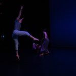 A dancer closest to the camera leaps in the air, toes pointed and legs outstretch. A dancer upstage of her is running, while another rolls on the floor.