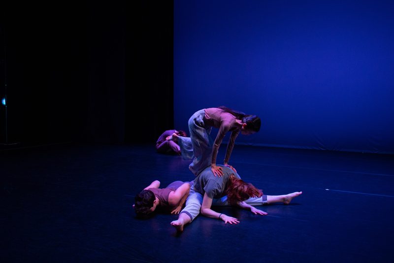 One dancer lies facedown on the stage. Another dancer is seated against her with legs outstretched, arms and face down. A third dancer has her hands on the second dancer's back, kicking her legs into the air.