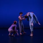 FourA dancer crouches on the stage and looks up to another dancer, who is standing straight up and helping a third dancer carry a fourth dancer, who is draped over her shoulders, stomach toward the ceiling.