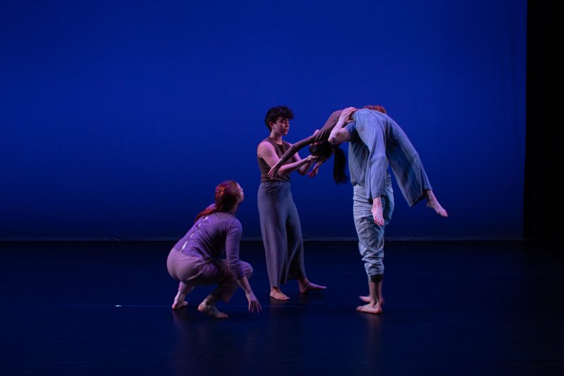 FourA dancer crouches on the stage and looks up to another dancer, who is standing straight up and helping a third dancer carry a fourth dancer, who is draped over her shoulders, stomach toward the ceiling.