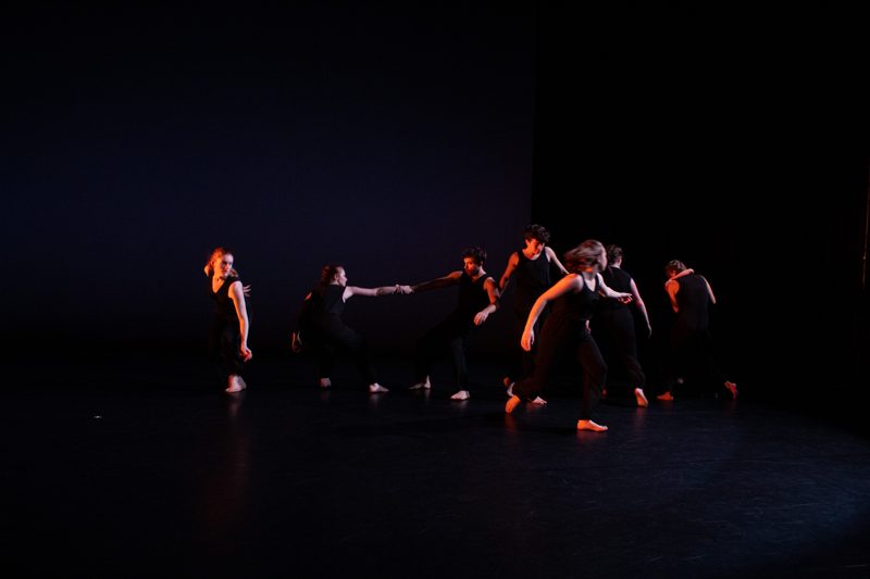 A group of dancers clothed in all black form a rough line across the stage. Some dancers are running, some pull at each other by the arm.