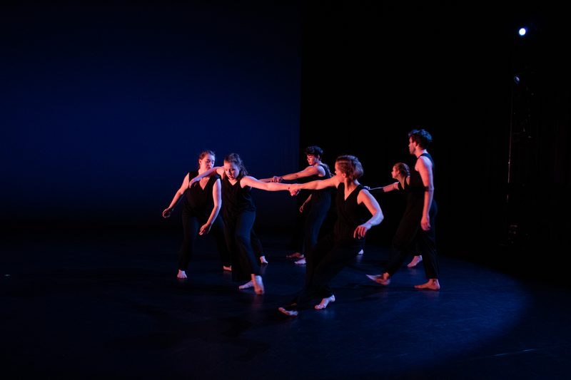 A group of dancers in black costumes stand and hold hands, pulling on each other and forming a rough circle.