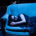 A person with black and white makeup across their face sleeps on a blue sofa under cool light.