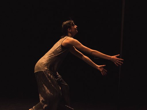 Side view of a dancer mid-movement, crouching down and reaching both arms toward stage left.