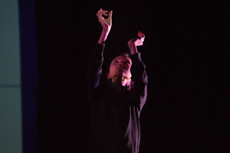 Front view of a dancer dressed in black, their face looking upwards, hands with wrists bent, reaching towards the ceiling.