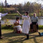 Three people, one of whom is playing a guitar, sit on crates and the ground while watching two people dressed in white looking at each other from six feet away. Their hands are stretched towards each other as though they are dancing.