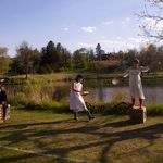 Three women in white dance with arms outstretched in front of a lake. A man dressed in black sits at a table made of a crate with a newspaper crown on his head, writing on a piece of paper.
