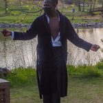 A man wearing an untied black robe and an unbuttoned shirt stands in front of a lake with his arms outstretched. His gaze is directed off to the left, where the corner of a crate is barely visible.