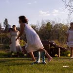 A woman in a white dress sprints to the left just behind a pair of blue rain boots. Two other women in white dresses stand on either side of a pile of crates and stare at the first woman. All actors are outside in a meadow with fake flowers planted in the grass.