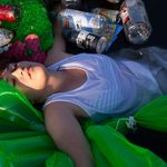 A woman wearing a white dress floats on her back in the water, surrounded by black plastic netting, plastic bottles, cans, and inflatable neon green bags. Her eyes are mostly closed and she has no expression on her face.