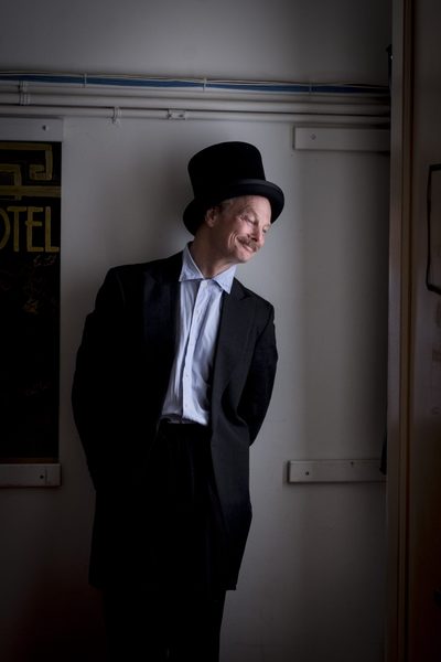 A person leaning with their back against a wall in a dimly lit room, wearing a black suit and black top hat. Smiling at the ground