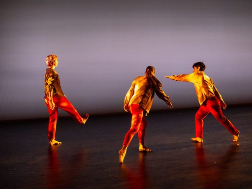 a black stage floor his a grey background, 3 dancers in various poses wearing red pants and yellow tops