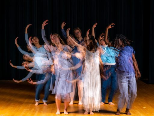A blurred image of about seven dancers dressed in various cool-toned colors have one arm raised above their head and are leaning over.