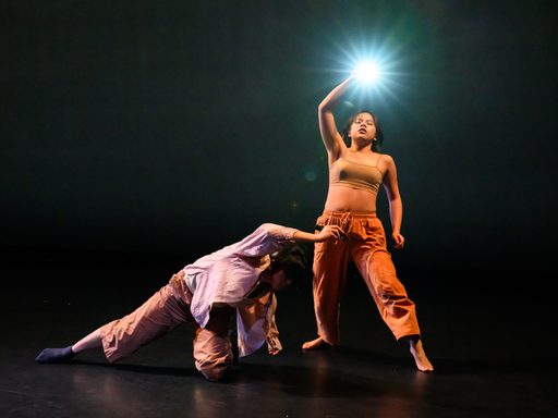 Two dancers on stage, one crouched with an arm extended, the other standing, reaching up with a bright light in hand.