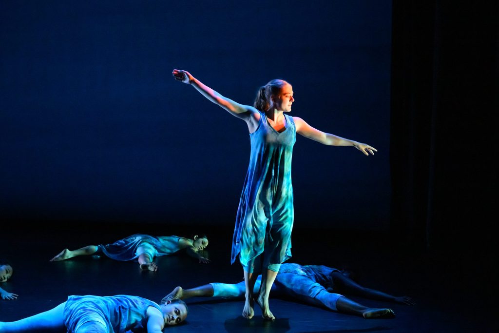 A dancer in a flowy blue dress stands gracefully with outstretched arms on a dimly lit stage, surrounded by other dancers lying on the floor.