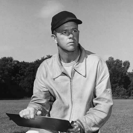 Football coach Warren Beson kneels on the grass holding a clipboard