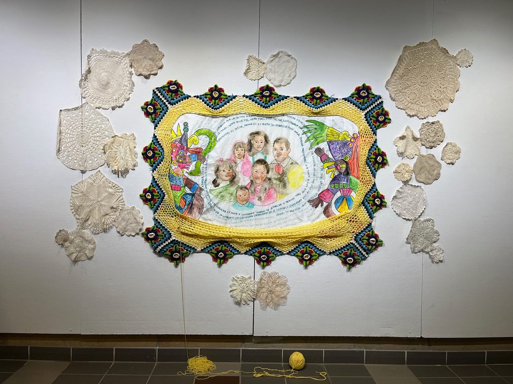 A painted family portrait surrounded by circular text and colorful images. The artwork is made from yarn, thread, muslin, acrylic paint, marker, and doilies