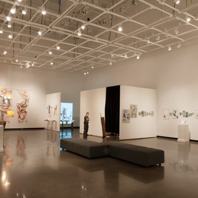 A brightly-lit art gallery with white walls. Some notable pieces include artificial flowers hanging from the ceiling to the right and a large sheet painted with bright colors hanging on the back wall.