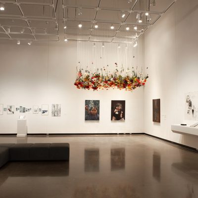 A brightly-lit art gallery with white walls. In the center of the room, hundreds of artificial flowers hang from the ceiling, while paintings and drawings hang on the walls.