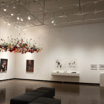 A brightly-lit art gallery with white walls. In the center of the room, hundreds of artificial flowers hang from the ceiling, while paintings and drawings hang on the walls.